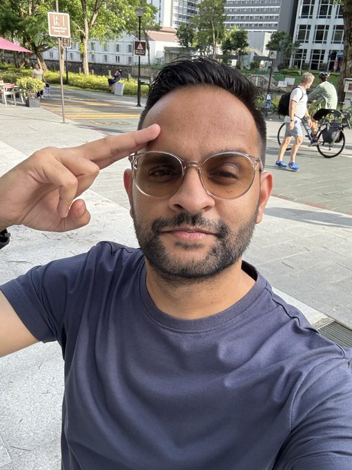 A man wearing tinted glasses and a navy shirt takes a selfie on a city street, holding two fingers up to his forehead.