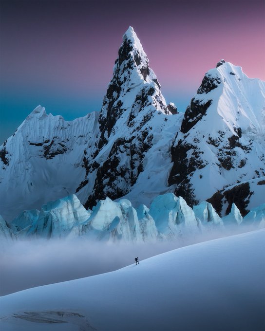 A dramatic landscape featuring a jagged, snow-covered mountain peak under a purple and teal gradient sky, with a small figure of a skier on a snowy slope in the foreground.