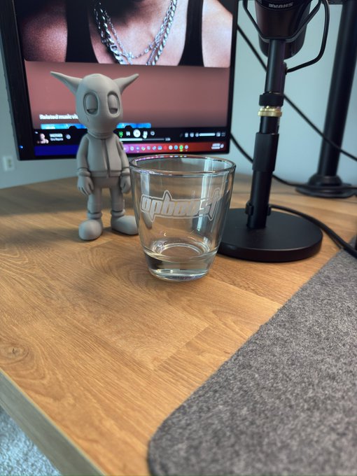 A small Astro Boy figurine stands next to a clear 'apbarfly' shot glass on a wooden desk, with a microphone and computer monitor in the background.