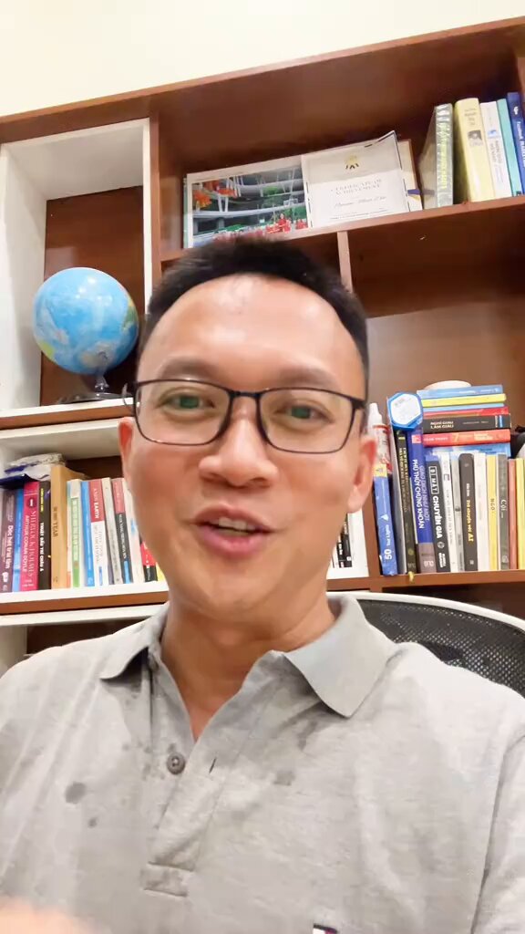 The image features a man wearing glasses sitting at a desk with numerous books surrounding him. The man appears to be talking into a microphone or camera while surrounded by an array of books, suggesting that he might be discussing a topic related to literature or research. The presence of multiple books in various sizes and colors indicates that the man is likely engaged in a discussion about reading materials or possibly even a book review.