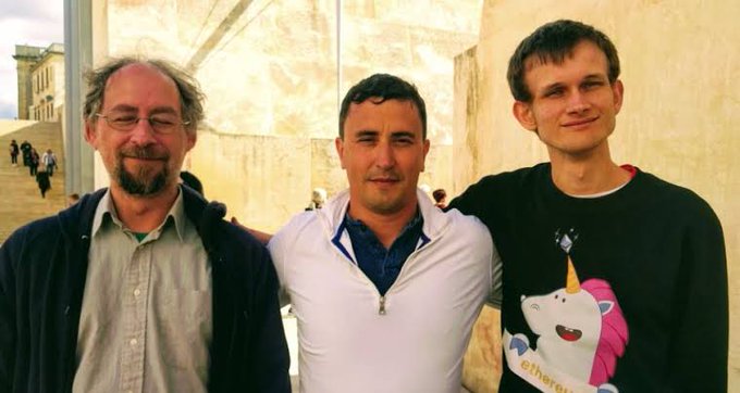 Three men pose together for a photo; the man on the right wears a black sweater featuring a cartoon character wearing a unicorn hat with the word 'ethereum' visible.