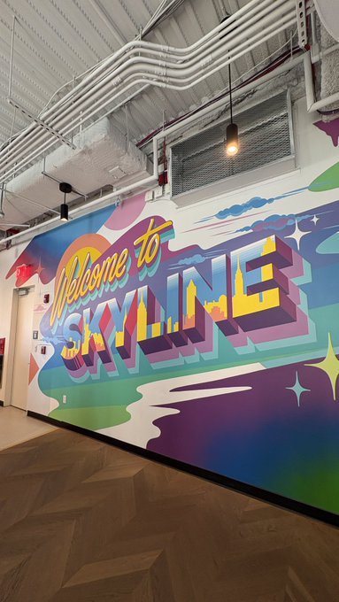 A colorful, retro-styled mural on an industrial wall reads 'Welcome to SKYLINE' in stylized lettering with a cityscape silhouette, surrounded by stars and abstract shapes.