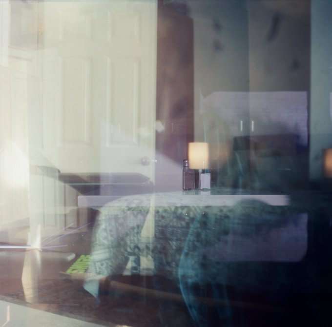 A blurry, dreamlike image showing reflections on glass, revealing a bedroom interior with a bed, lamp, and pillows.