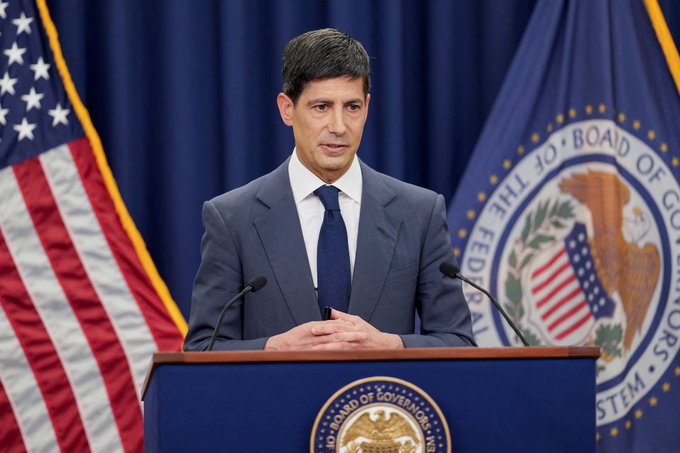A man in a gray suit speaks at a podium flanked by the American flag and the Federal Reserve seal, addressing an audience.