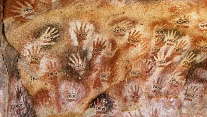 Ancient cave art featuring numerous handprints and eye-like symbols painted in red and black on a textured rock wall.