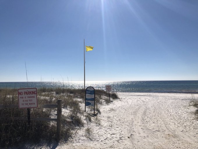 A yellow flag flutters over a sunlit beach path, with a red sign reading "NO PARKING PUBLIC PARKING IS ALSO TO THE NORTH" visible on the left. The "GM" caption likely refers to the hazard-warning yellow flag typically used on Gulf Coast beaches, such as at Gulf Shores or Pensacola, to indicate moderate surf or marine hazards.

**Text visible:** "NO PARKING / PUBLIC PARKING / IS ALSO / TO THE NORTH"
