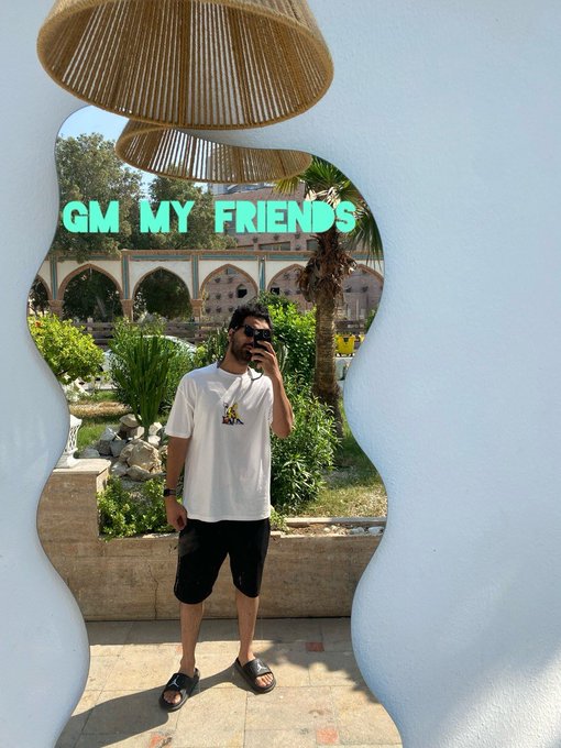 A man in a white t-shirt and black shorts takes a selfie in a wavy, blue-framed mirror with the text 'GM MY FRIENDS' displayed above him, reflecting a garden with arches and palm trees.