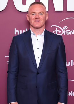 A man in a navy blue suit and light blue dress shirt poses on a red carpet at a Hulu and Disney event. He has short dark hair and is smiling at the camera.