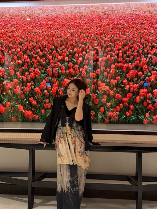 A woman in a black blazer and patterned dress poses in front of a large illuminated display of a red tulip field.