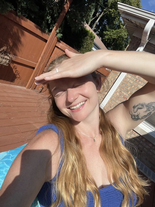 A young woman with long blonde hair smiles brightly while shielding her eyes from the sun with her hand, wearing a blue top and showing a tattoo on her upper arm. The sunny outdoor setting near a wooden fence complements the cheerful "Good morning 🌞" vibe of her post.