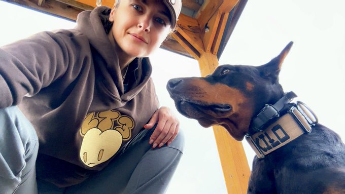 A young person wearing a brown BAPE hoodie and cap sitting under a wooden structure, looking at a Doberman dog wearing a tactical collar.