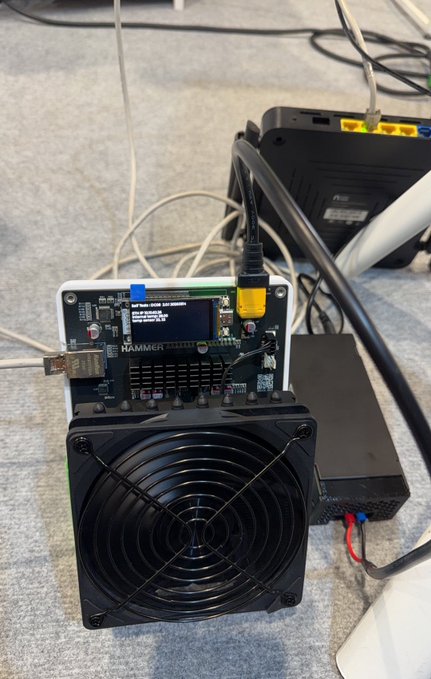 A Bitcoin mining setup featuring a HAMMER brand ASIC miner with a cooling fan, connected to a network cable and power supply, sitting on a carpeted floor.