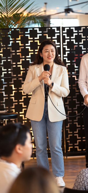 A woman in a beige blazer and jeans holds a microphone while speaking, smiling at an audience in front of a decorative gold geometric room divider.