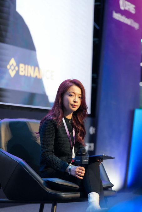 Photo of a woman with reddish-brown hair seated on a panel at what appears to be a Binance Institutional Forum event, with a Binance logo visible in the background.