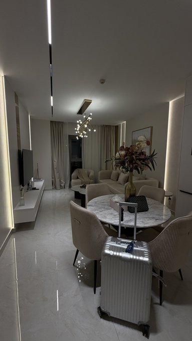 A dimly lit, modern hotel or apartment living area with a marble dining table, a beige sofa, a decorative chandelier, and a silver suitcase placed in the foreground.