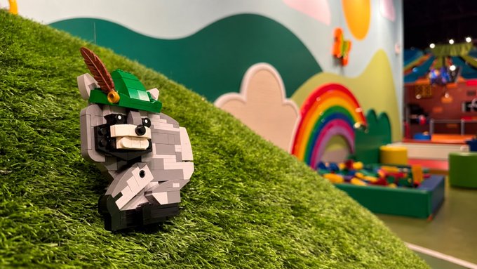 A LEGO figure of a raccoon-like character (Jimothy) wearing a green Robin Hood hat with a feather sits on artificial green turf, while in the background a colorful mural with a rainbow and a bin of scattered LEGO bricks is visible inside what appears to be an indoor play area.