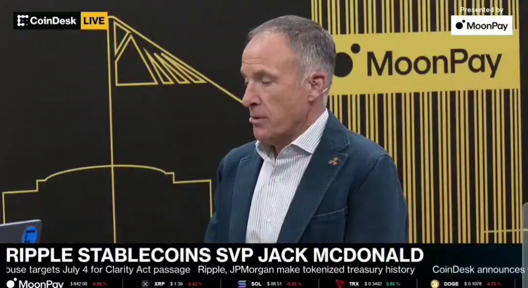 The image shows a man wearing a suit, standing next to a computer screen displaying an advertisement for Ripple. The advertisement features a man talking about Bitcoin and its potential uses. The text on the screen reads "Ripple Stablecoins SVP Jack McDonald", which is related to the cryptocurrency Ripple. Additionally, there are mentions of other cryptocurrencies like Bitcoin and Litecoin in the image.