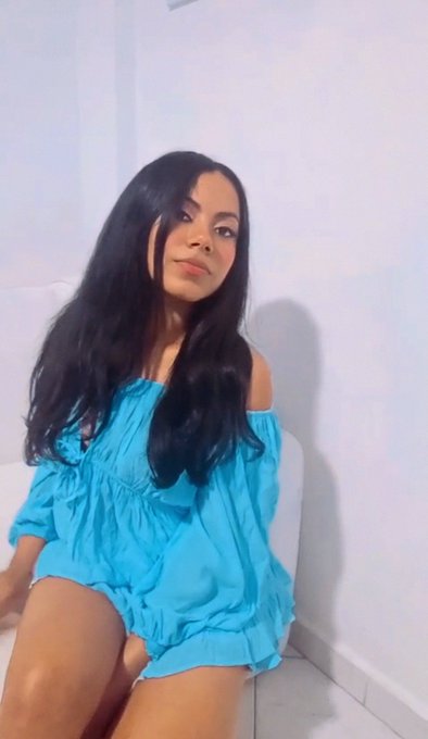 A woman with long dark hair wearing a blue off-the-shoulder romper sits on a white couch against a plain wall.