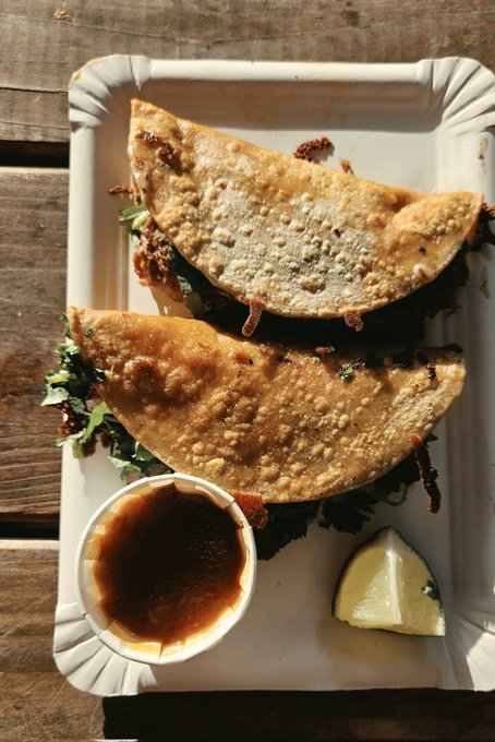 An overhead photo of two folded crispy tacos on a white paper tray, served with a small cup of dark sauce and a lemon wedge on a wooden table.
