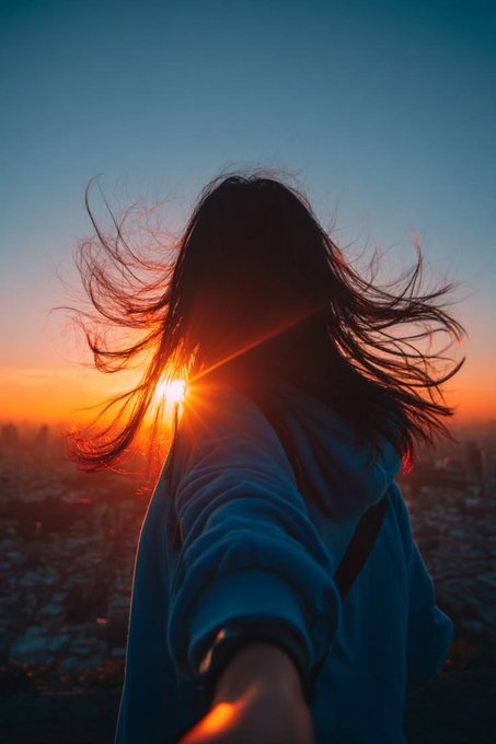 Silhouette of a person with flowing hair against a vivid orange and blue sunset, with cityscape visible below.