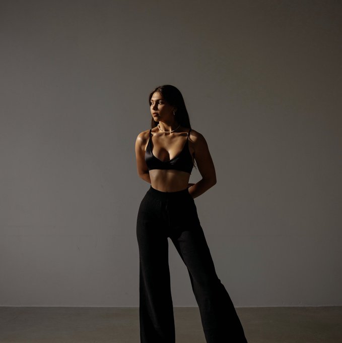 A young woman with long dark hair poses in a studio wearing a black halter bra top and high-waisted black flared pants, lit dramatically against a gray backdrop.