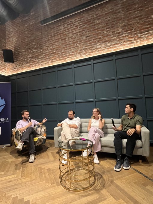 Four people are seated in a panel discussion setting in a room with brick walls and green paneled walls, holding microphones, with one person on a couch and another in an armchair.