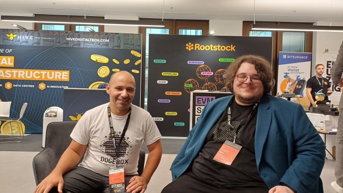 Two men sit smiling at a conference booth with banners for Hive Digital Tech and Rootstock visible in the background; one wears a 'Doge Box' t-shirt.