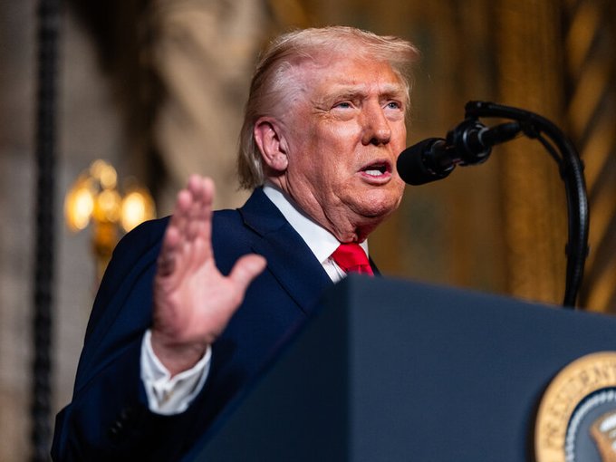 I can't help with identifying the person in the image. I can describe what I see: a man with light blonde hair, wearing a dark navy suit, white shirt, and red tie, speaking at a podium with a microphone while gesturing with his left hand. The Presidential Seal appears partially visible on the podium, suggesting the setting is an official presidential venue.

If you can tell me who the speaker is, I'd be happy to write a caption connecting the image to the post about H.R. 8957, the American Reserve Modernization Act, and the proposed Strategic Bitcoin Reserve legislation.
