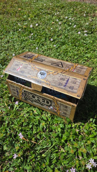 A small decorative treasure chest with "WANTED" posters and the word "GOLD" printed on it sits in a grassy field dotted with small flowers.