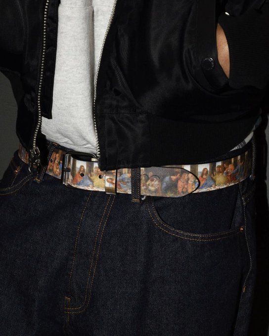 A close-up photograph of a person wearing a black jacket, gray shirt, and dark jeans, fastened with a distinctive belt featuring a classical Renaissance-style religious painting design.