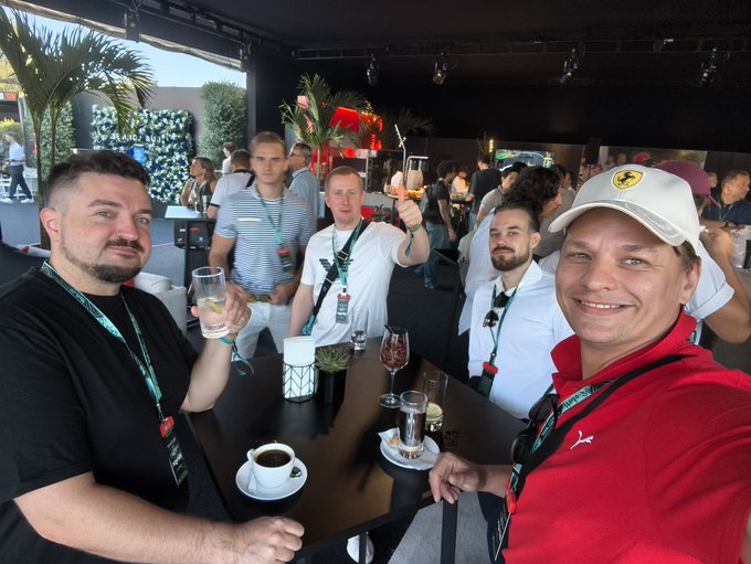 A group of five men pose together at an outdoor venue, smiling and toasting with drinks; one wears a red shirt and a white Ferrari cap.
