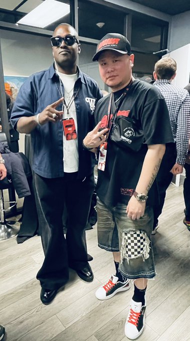 Two stylish individuals pose together indoors, with the man on the right wearing a black "Bored Ape" branded cap, matching graphic tee, and checkered shorts, while sporting "Big Poppa" sneakers. The man on the left, identified as @illaDaProducer, complements the look with sunglasses and a blue BAYC overshirt, fitting the caption's celebration of two "best dressed apes in the club."