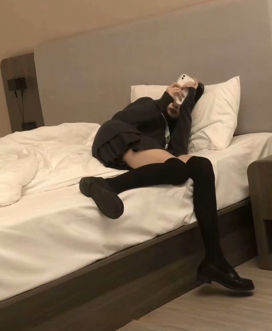 The image features a person laying down on their bed, holding a cell phone up to their face as if taking a selfie or capturing a photo of themselves. The person appears to be dressed in black clothing with knee-high socks. There are two clocks visible in the room, one near the top left corner and another towards the bottom right corner.