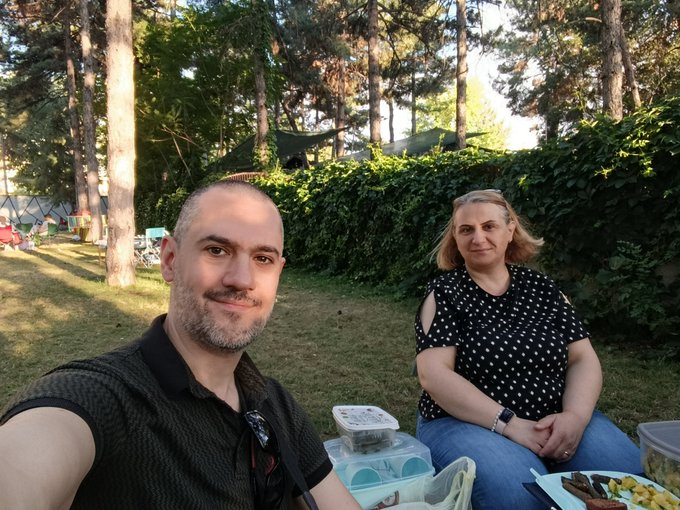 A man and woman take a selfie while enjoying a meal outdoors at a campsite surrounded by tall trees and a green hedge, capturing a relaxed "touching grass" moment in nature.