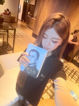 A woman with shoulder-length hair taking a mirror selfie in a restaurant while holding up a blue book with a man's face on the cover and Chinese text.