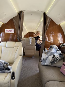 Interior view of a private jet cabin showing cream leather seats, wood paneling, and a woman in a dark dress standing near the back, with various bags and clothing items scattered on the seats.