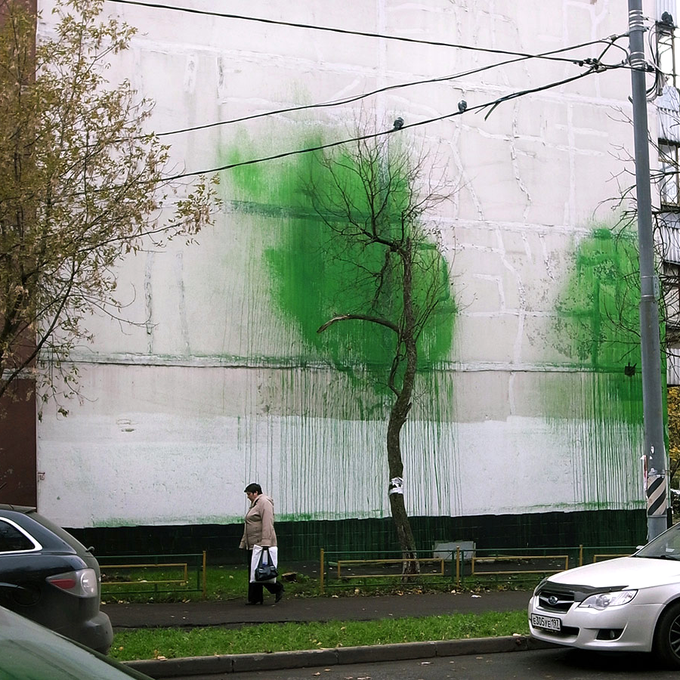 A large white building wall is covered in green spray paint graffiti, while an elderly woman carrying a bag walks along the sidewalk in the foreground next to a parked silver car.