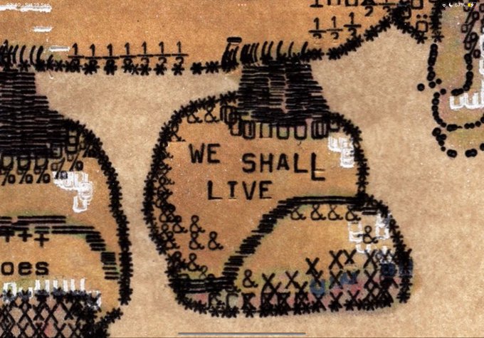A textured mixed-media artwork on a brown surface features the stenciled text "WE SHALL LIVE" surrounded by abstract designs made from characters, numbers, and symbols.