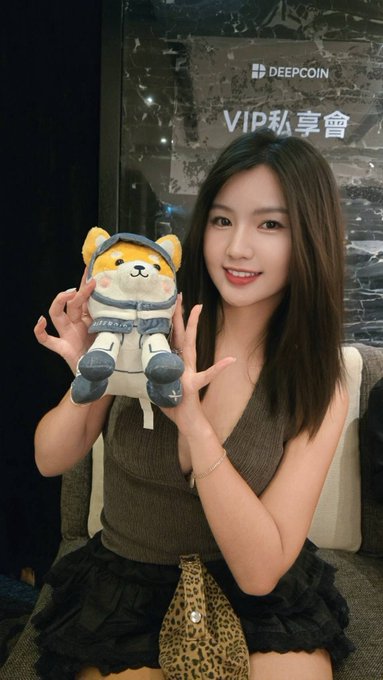 A young woman with long dark hair holds up a Shiba Inu plush toy wearing a spacesuit in front of a glass door with 'DEEPCOIN VIP 私享會' text.