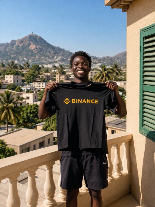 A smiling man stands on a balcony overlooking a tropical cityscape, proudly holding up a black t-shirt displaying the yellow "BINANCE" logo, proudly identifying as a "Binancian."