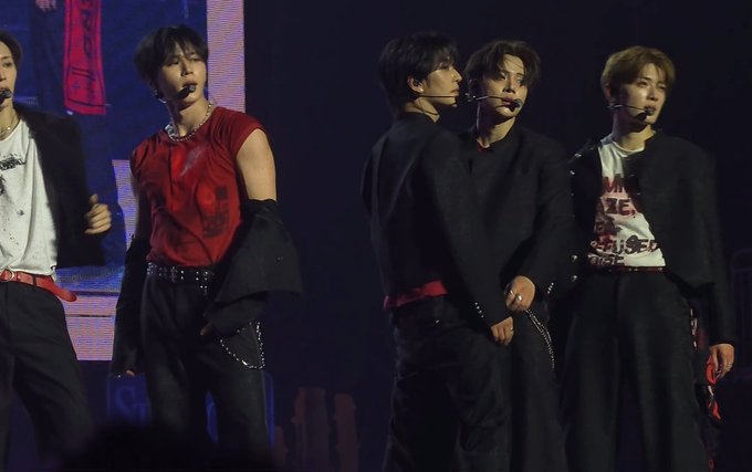 Concert photo of five male K-pop performers on stage, with one wearing a red sleeveless top and others in black outfits, all wearing headset microphones.