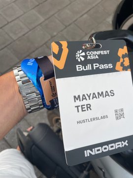 A hand wearing a metal watch and blue "Liquid" wristband holds a "Coinfest Asia Bull Pass" badge labeled "MAYAMAS TER" with "Hustlerslabs" and "IndoDax" branding, along with a QR code, suggesting the holder is attending the Coinfest Asia crypto conference with sponsorship from IndoDax and Hustlerslabs, tied to the X profile linked in the post.