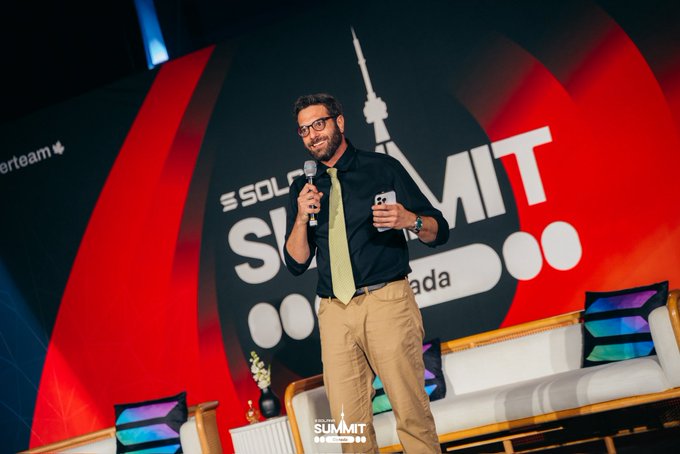 A man in a black shirt and yellow tie speaks into a microphone on stage at the Solana Summit in Toronto, with the event's logo prominently displayed on the backdrop behind him. Holding a phone, he engages with the audience at the Superteam Canada-hosted event, embodying the MC's reflection on public speaking as practice that builds confidence over time.
