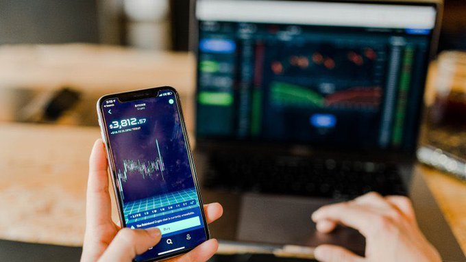 A person holds a smartphone displaying Robinhood's app showing a balance of $3,812.57, with a laptop displaying trading charts visible in the background.