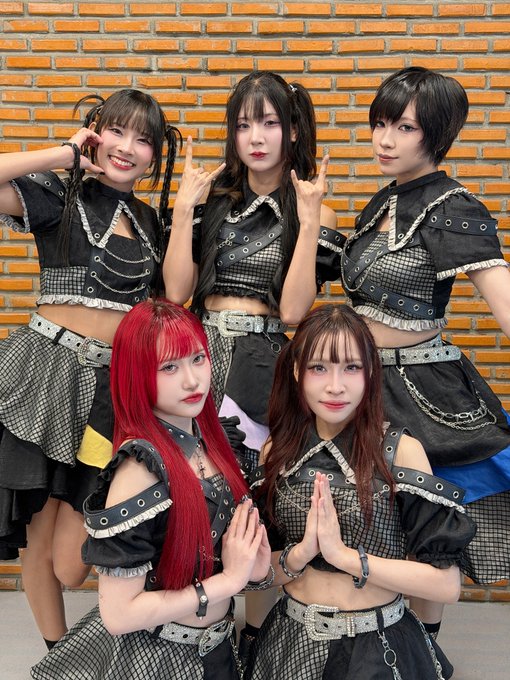 A photo of five young women dressed in matching black and grey plaid gothic-style idol outfits with chains and grommets, posing together with various hand gestures in front of a brick wall.