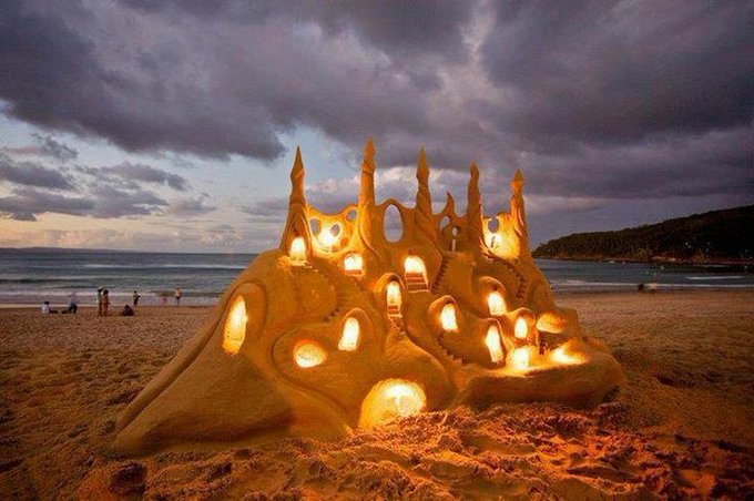 A sandcastle illuminated from within by candles stands on a beach at dusk, its elaborate, fragile architecture symbolizing the deceptive allure of crypto projects—much like the 100M+ tokens created, the structure may appear grand and inviting, but is essentially built on sand and destined to crumble, warning investors to be wary of who they follow in a market flooded with scams.