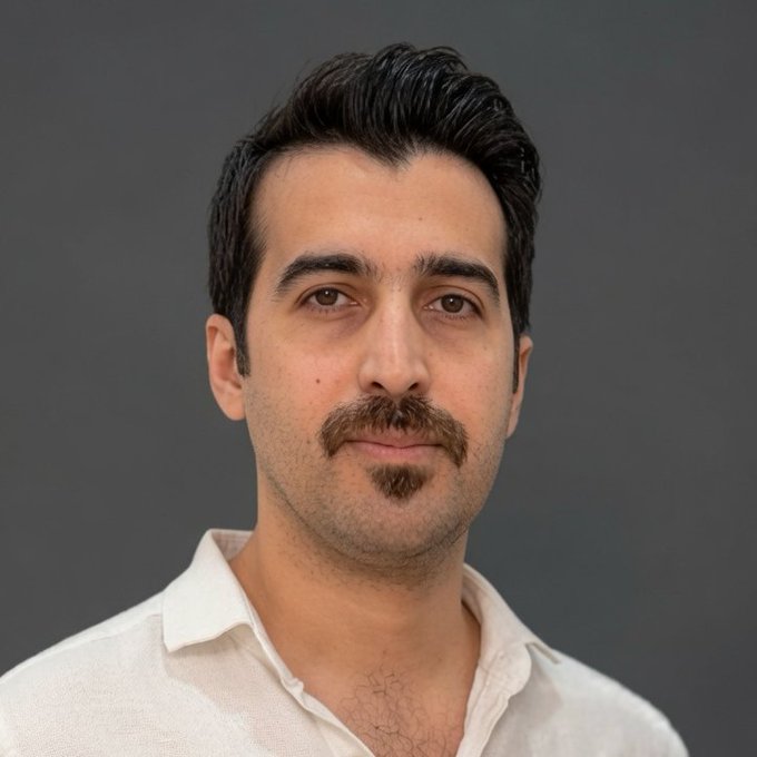 A studio portrait of a man with dark hair, a mustache, and a goatee, wearing a white collared shirt against a gray background.
