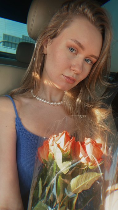 A portrait of a young woman with long blonde hair wearing a blue top and pearl necklace, holding a bouquet of orange and white roses in a car.