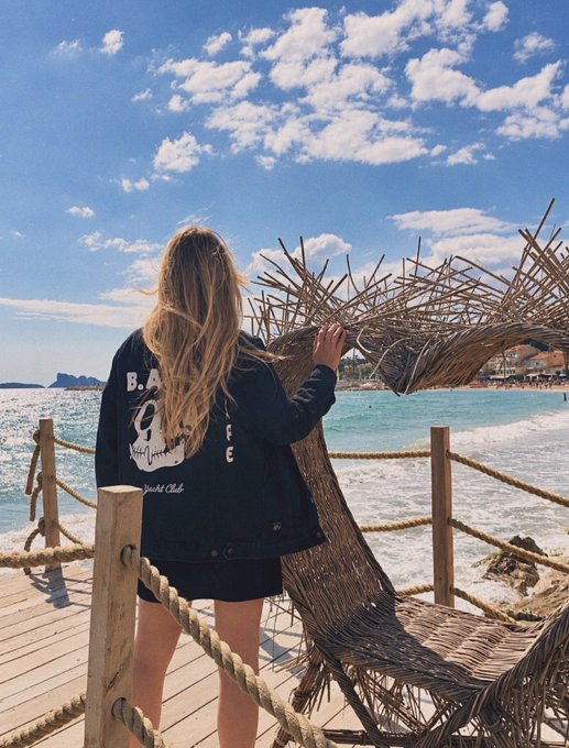 A woman with long blonde hair stands on a wooden pier overlooking a turquoise sea, wearing a black denim jacket featuring the "B.A.Y.C" text and a skull graphic, alongside a large woven sculpture of a sea creature. The jacket's bold lettering ties into the post's "B.A.Y.C" theme, complementing the breezy coastal backdrop and artistic seaside installation.
