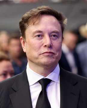 A photograph of Elon Musk wearing a black suit and tie, looking slightly to the side with a neutral expression against a blurred background.
